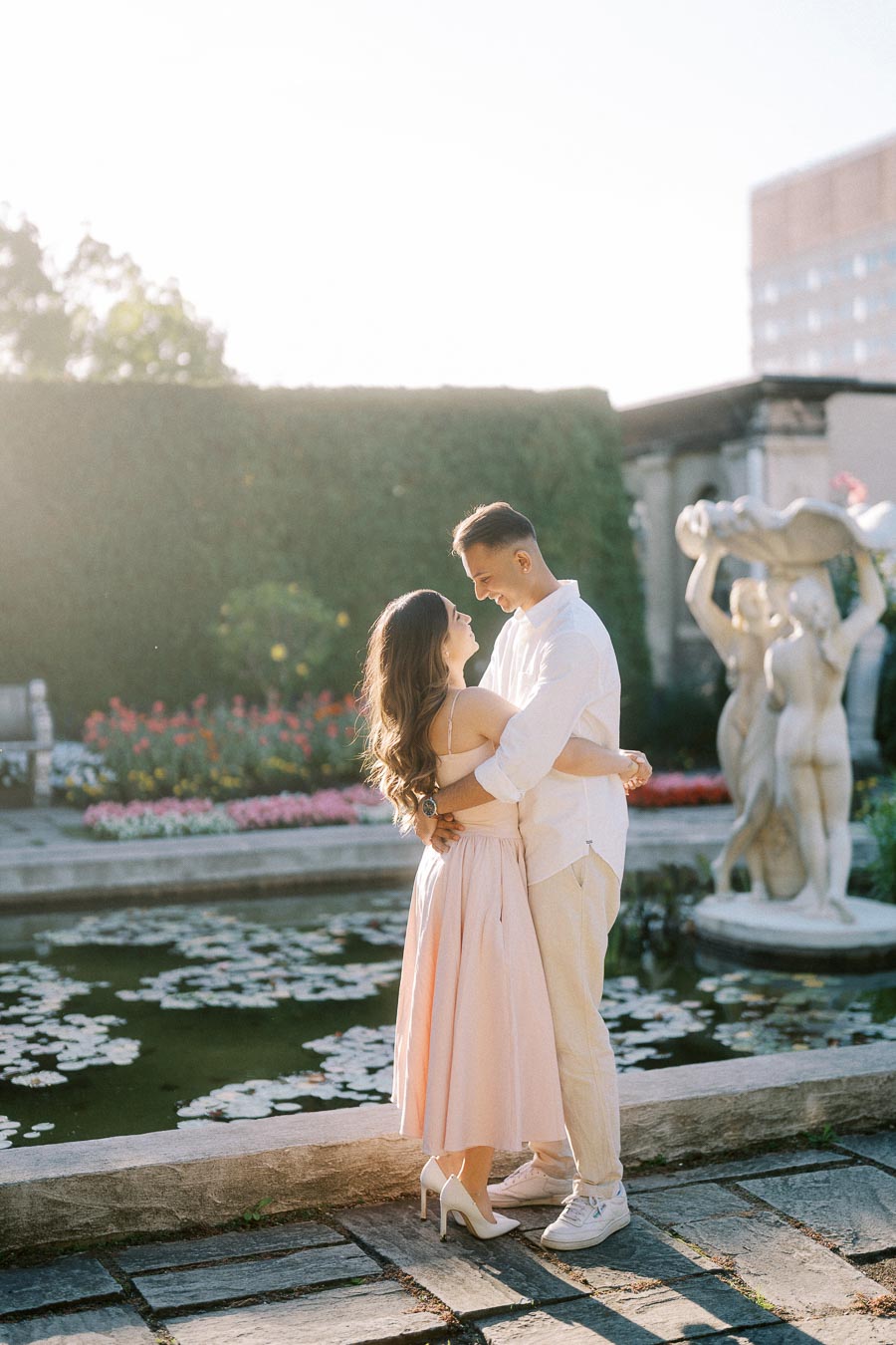 A couple embracing in a garden setting, surrounded by flowers and a lily pond, with sunlight softly illuminating their figures.