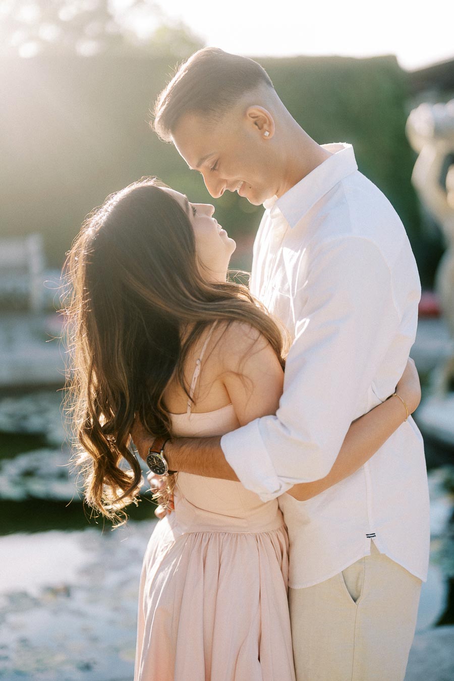 Young couple embracing outdoors on a sunny day, both smiling and wearing light summer clothing in front of a blurred garden backdrop.