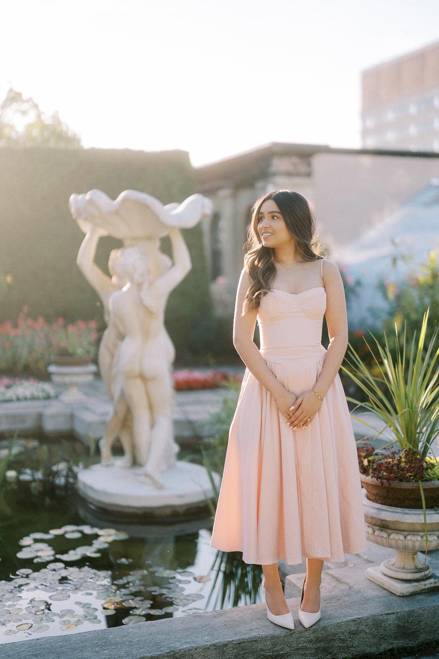 A woman in a light pink dress stands by a garden pond with a decorative fountain statue in the background, surrounded by lush greenery and flowers under soft sunlight.