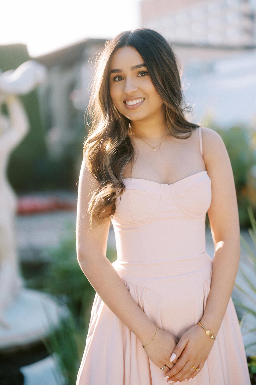 Smiling woman in a pale pink dress posing outdoors with natural light highlighting her hair, against a soft-focus background of greenery and a statue