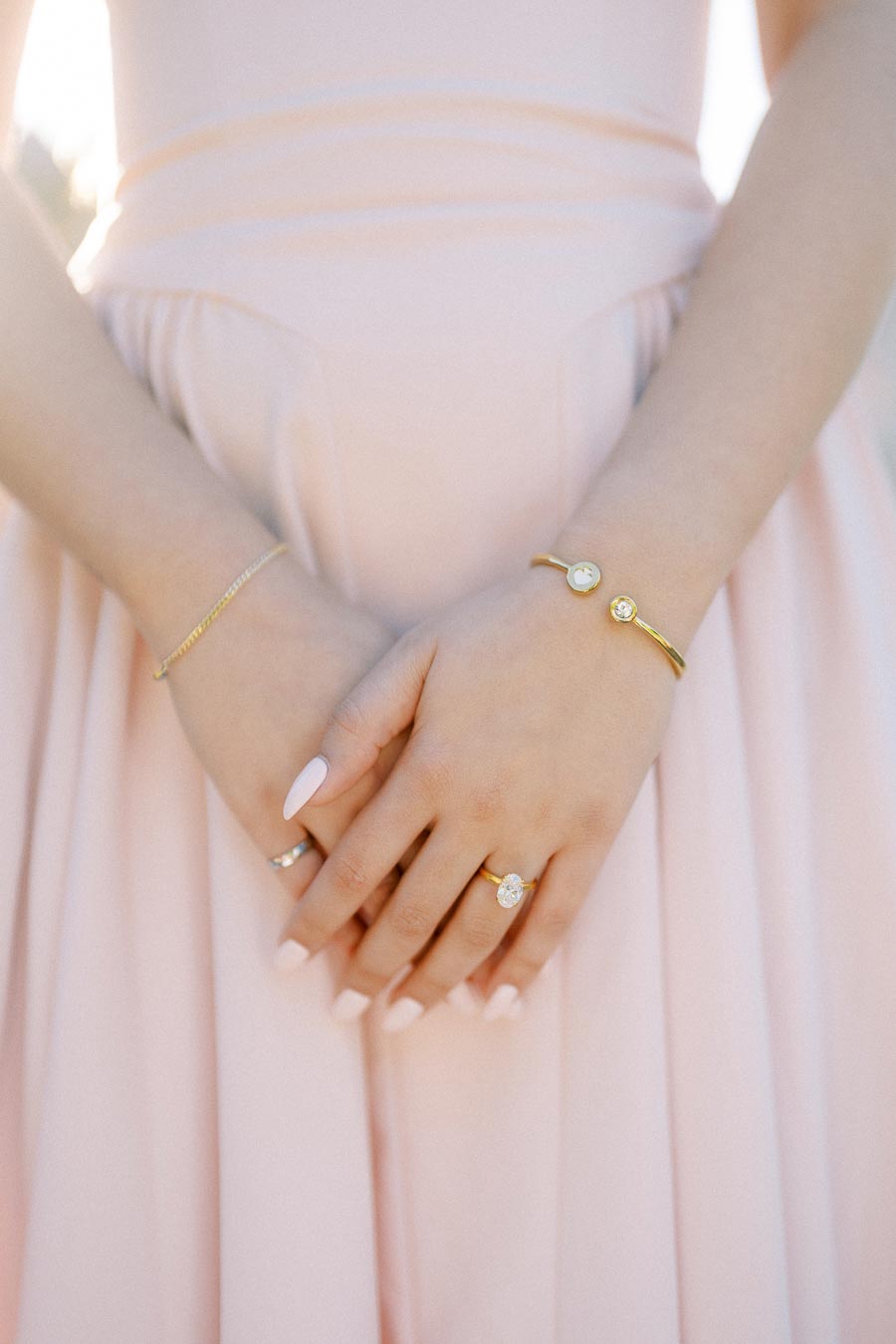 A woman in a light pink dress elegantly showing off her hands adorned with delicate gold jewelry, including bracelets and a ring, against a softly lit background.