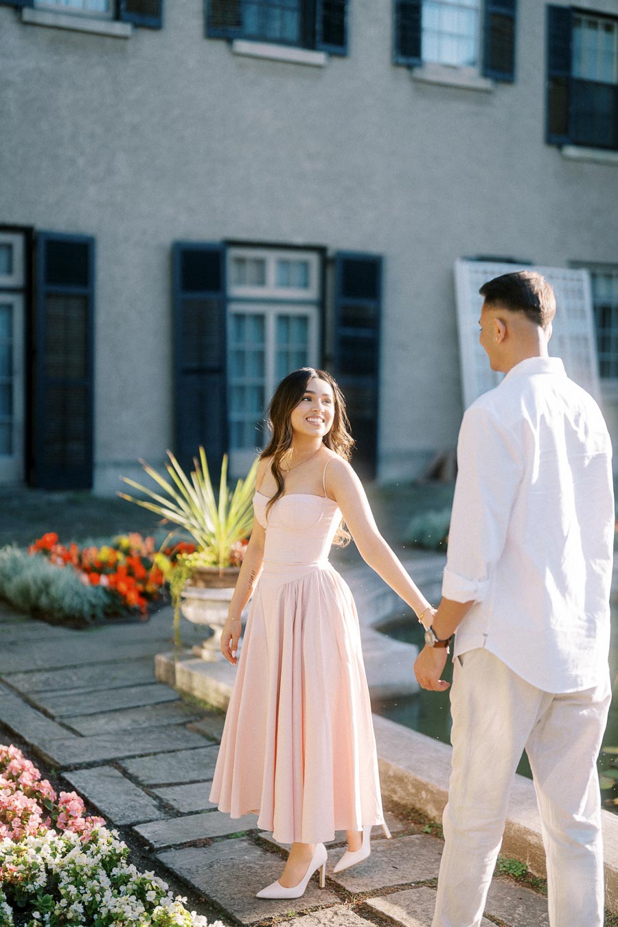 A couple enjoys a sunny day in front of a historic building, with the woman in a pink dress smiling warmly, holding hands with a man in white attire, surrounded by colorful flowers and greenery.