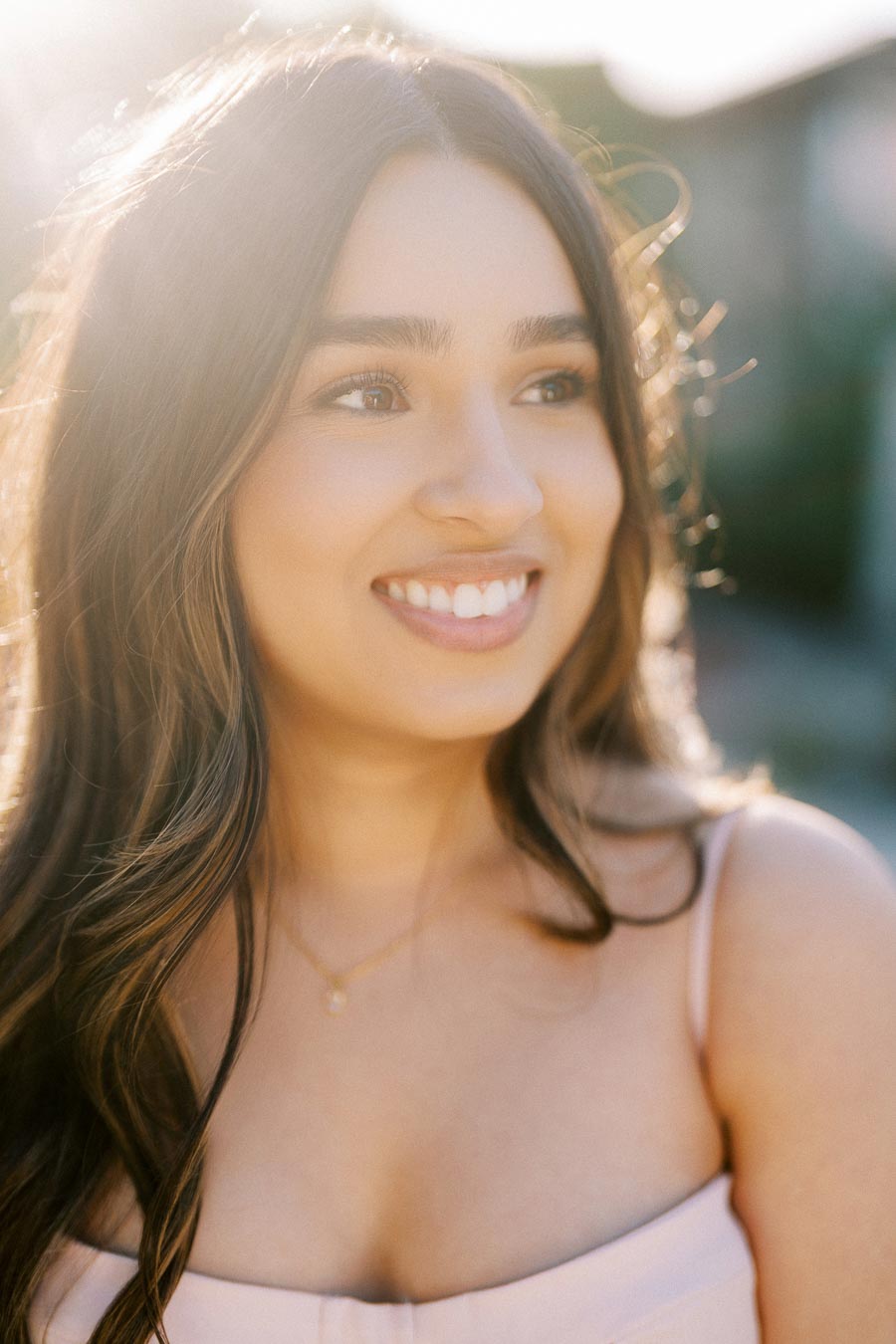 A woman smiling in sunlight, with long hair and a light summer outfit, enjoying an outdoor setting with a soft, natural background.