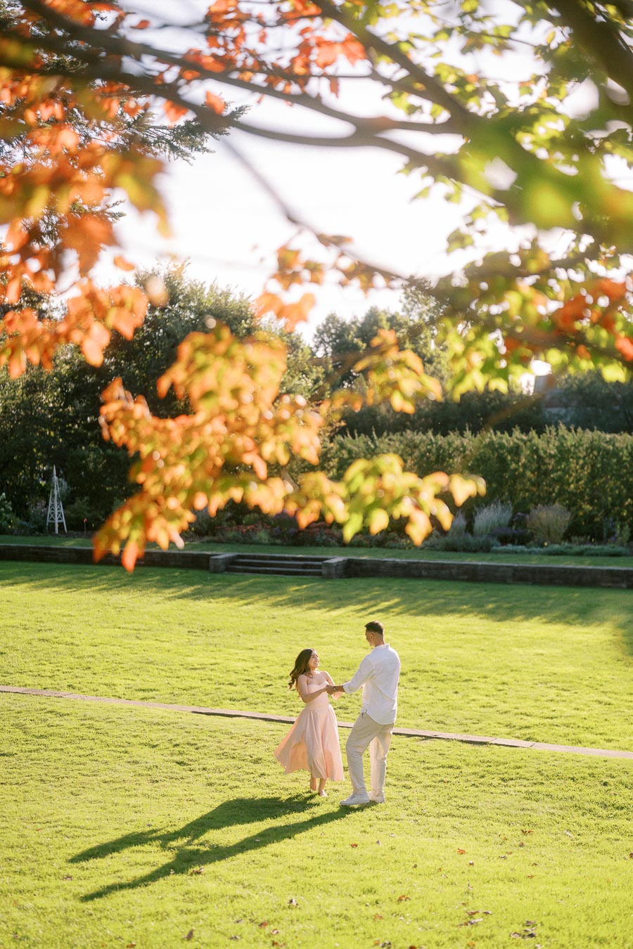 A couple dancing on a sunlit green lawn surrounded by vibrant autumn foliage, capturing a romantic moment in a picturesque park setting.