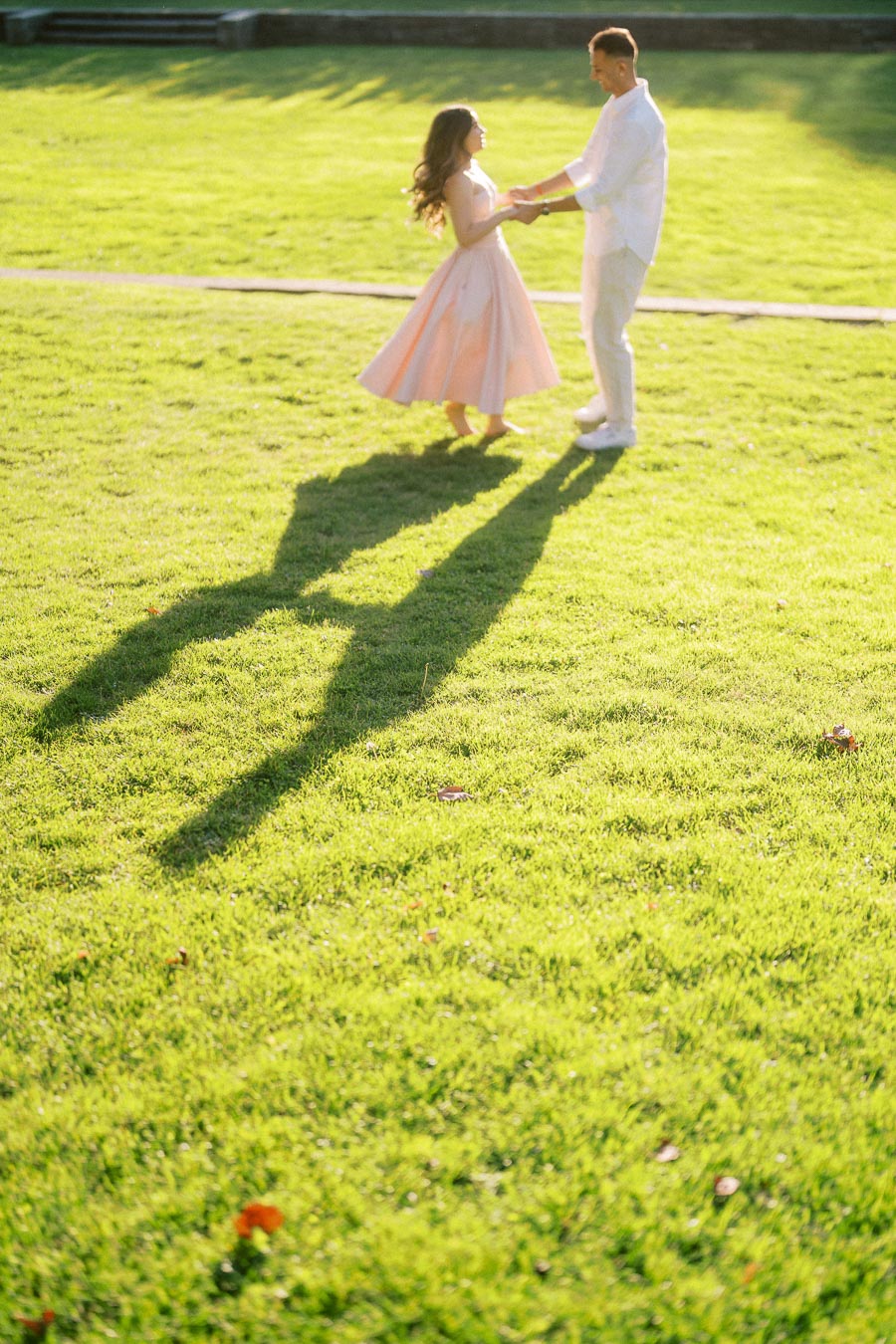 A couple dances gracefully on a sunlit grassy field casting long shadows, with the woman in a flowing pink dress and the man in a white outfit, evoking a romantic and joyful moment.