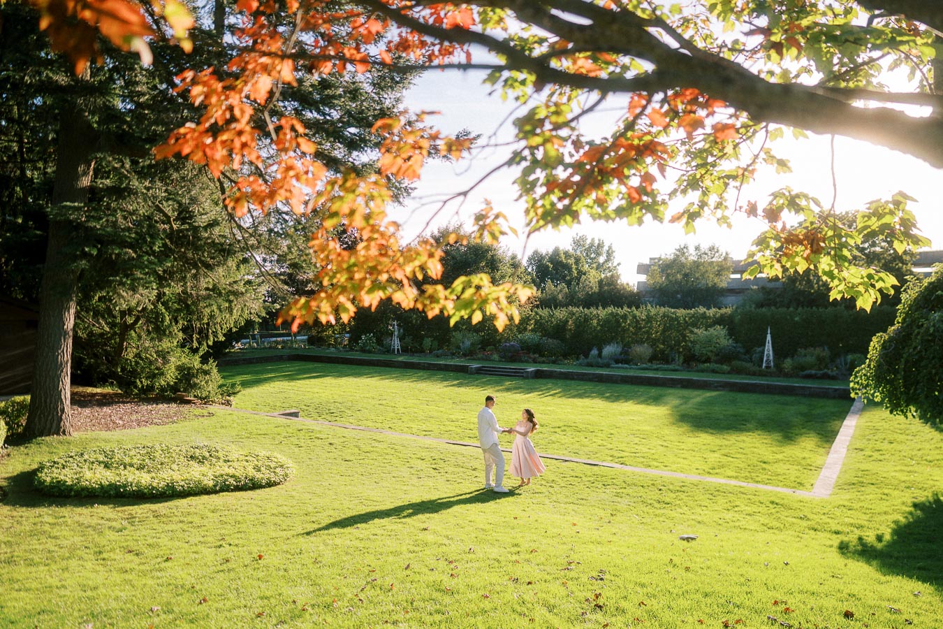 A couple dances on a sunlit grassy lawn surrounded by lush trees and a manicured garden, with autumn leaves in the foreground, creating a romantic outdoor setting.