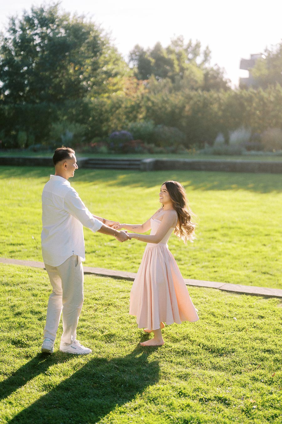 A couple smiling and holding hands while dancing on a sunny day in a lush green park.