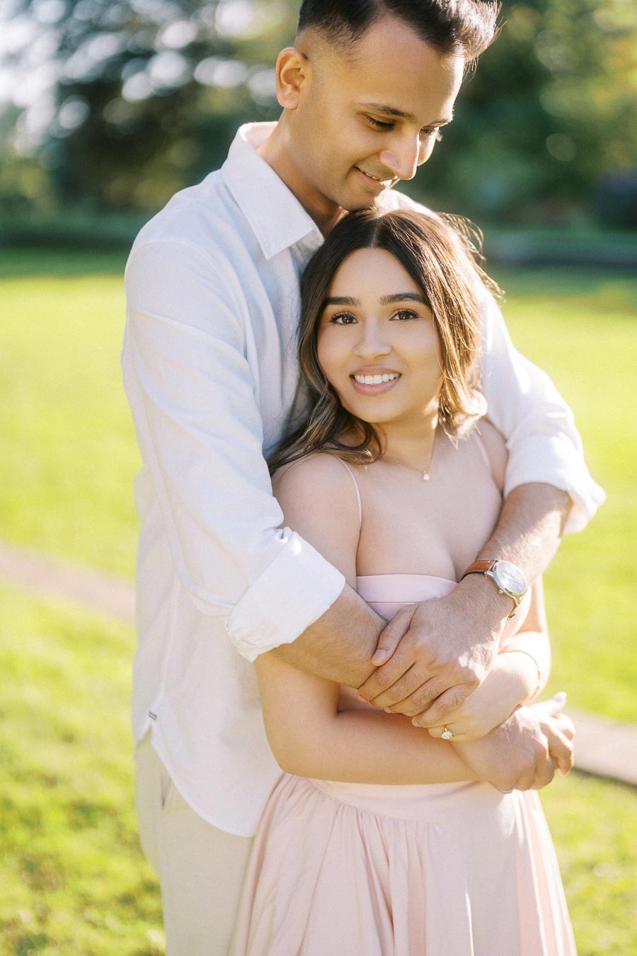 A couple embraces in a sunny park, the man gently holding the woman from behind, both smiling warmly. She's wearing a light pink sleeveless dress, and he's in a white shirt. The lush green background suggests a serene, outdoor setting.