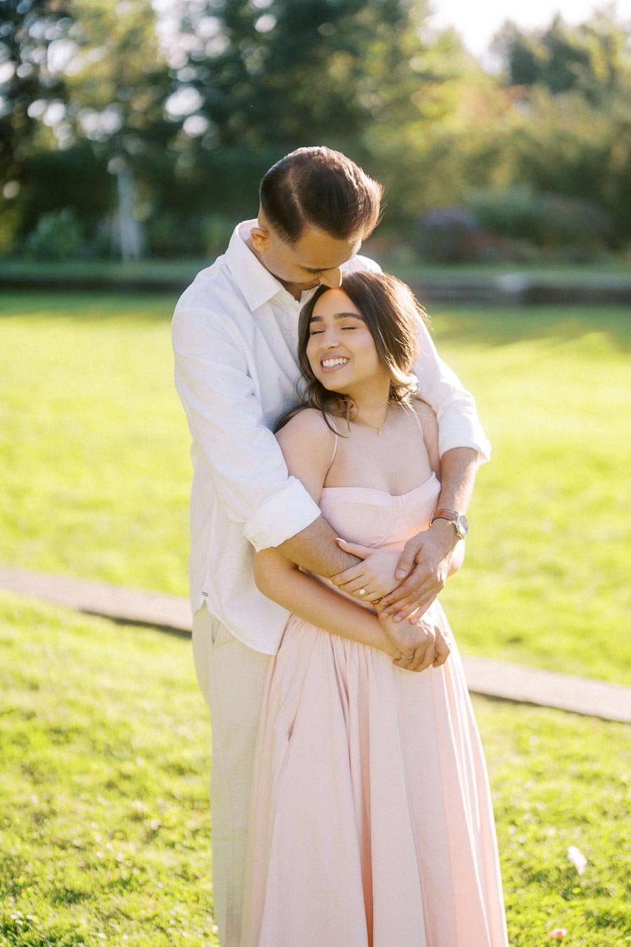 A couple embracing in a sunlit park, with the woman wearing a light pink dress and the man in a white shirt, exuding happiness and romance.