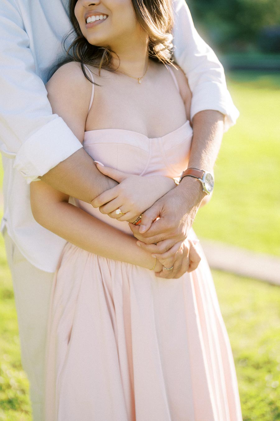 A couple embracing in a sunny park, with the woman wearing a light pink dress and the man in a white shirt, showing a close-up of their intertwined hands and the woman's joyful smile.