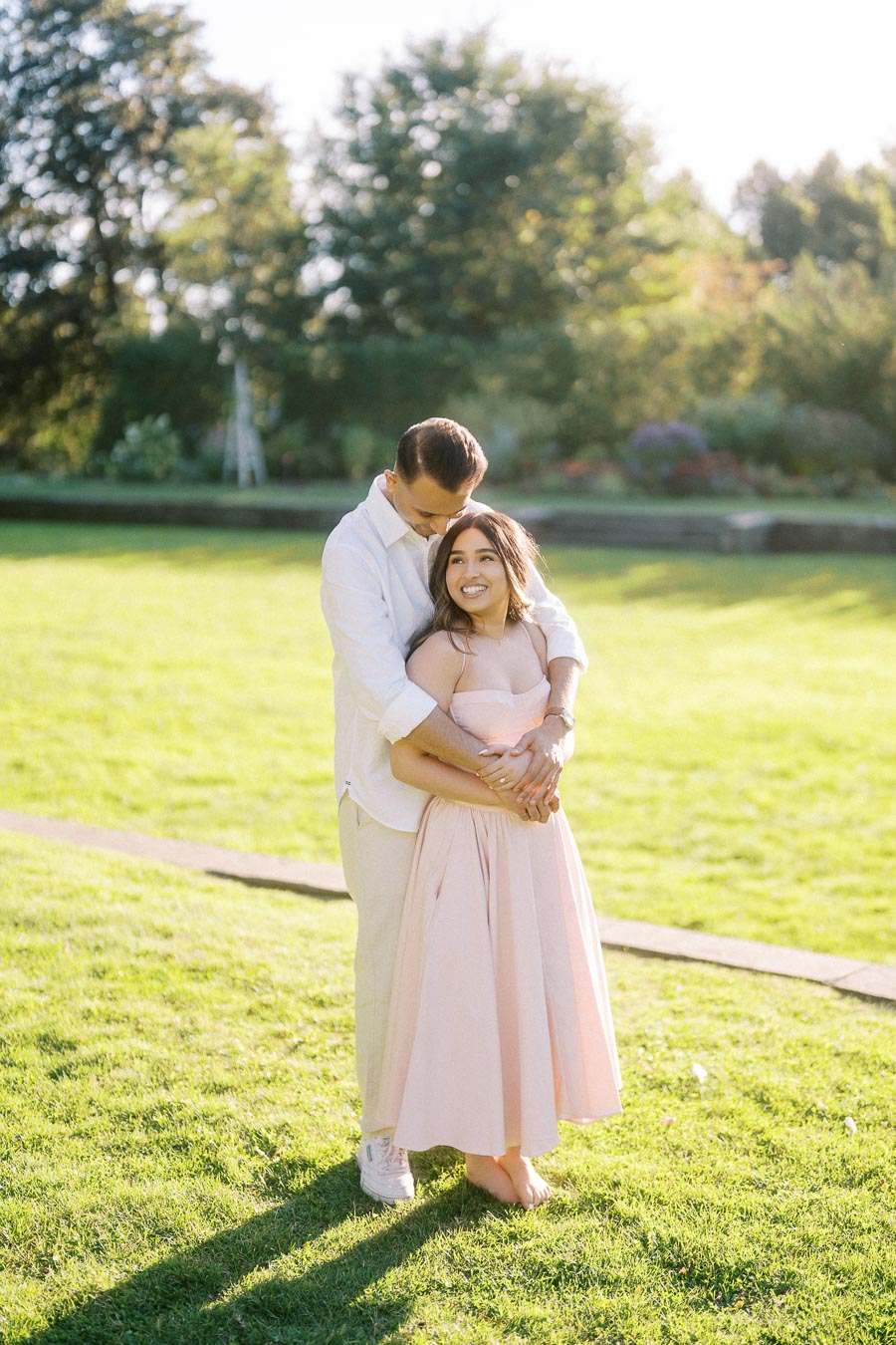 A couple embracing lovingly in a sunlit park, with a woman in a light pink dress and a man in a white shirt, surrounded by lush green grass and trees in the background.