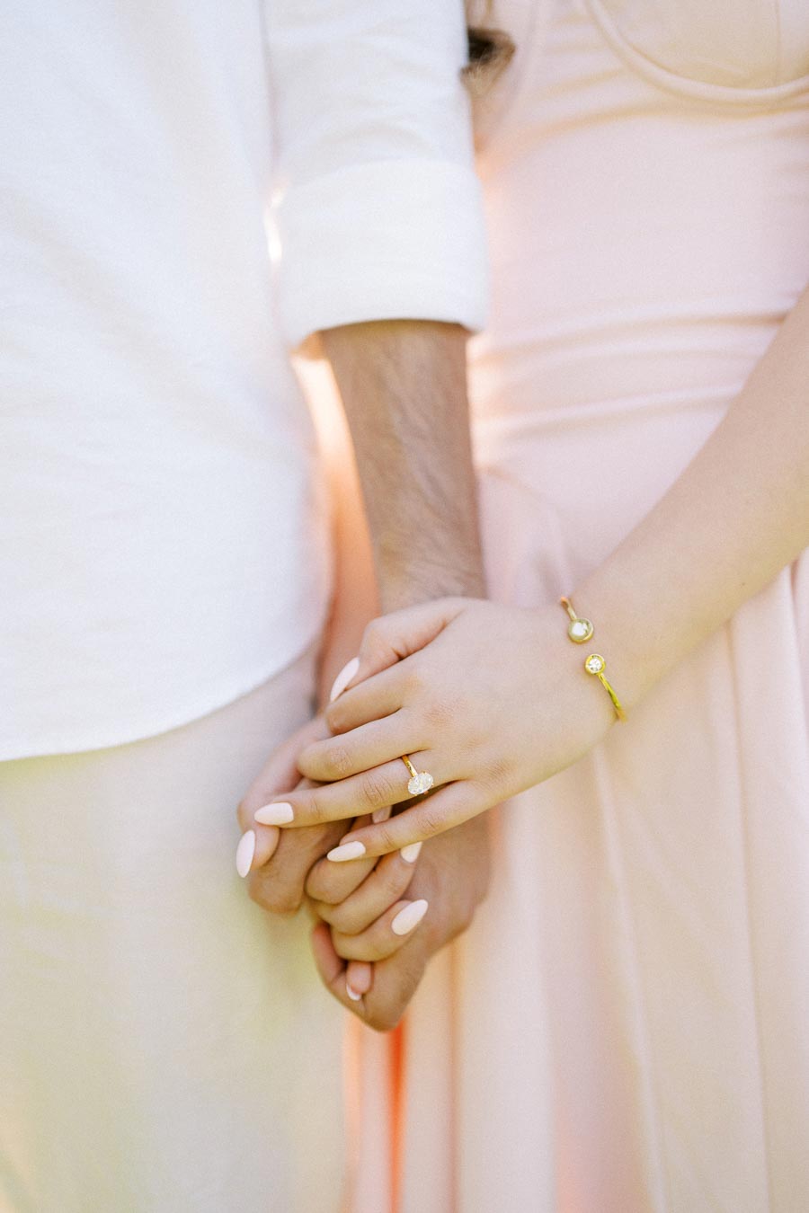 A couple holding hands, highlighting a diamond engagement ring and gold bracelet, dressed in soft pastel clothing.