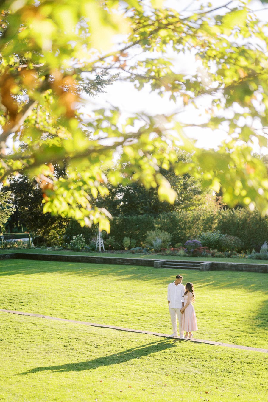 A couple standing on a sunlit path in a lush green garden, surrounded by vibrant foliage and soft afternoon light.