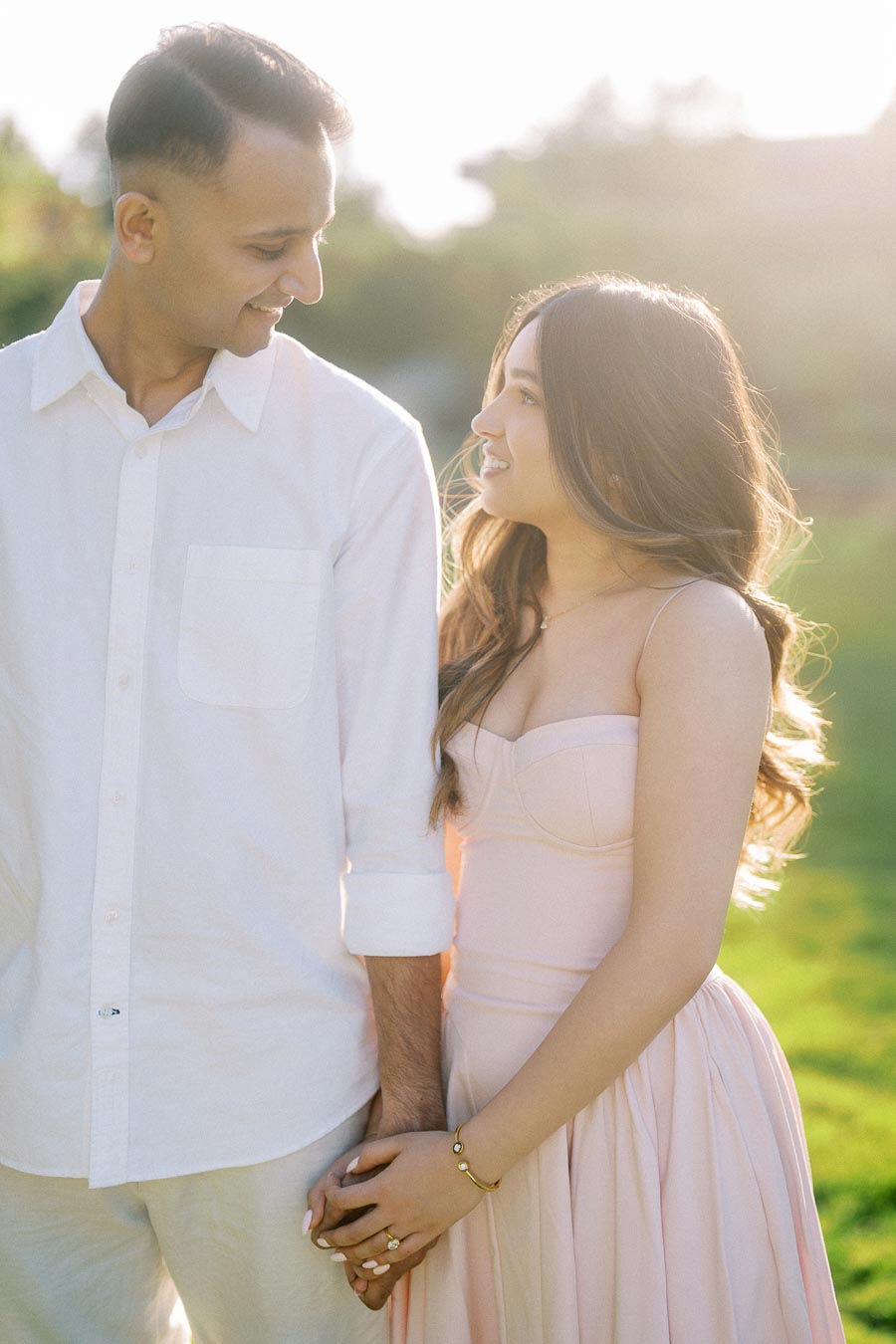 A couple standing closely in a sunlit garden, looking at each other affectionately. The woman is in a pastel pink dress, and the man is wearing a white shirt, both holding hands.