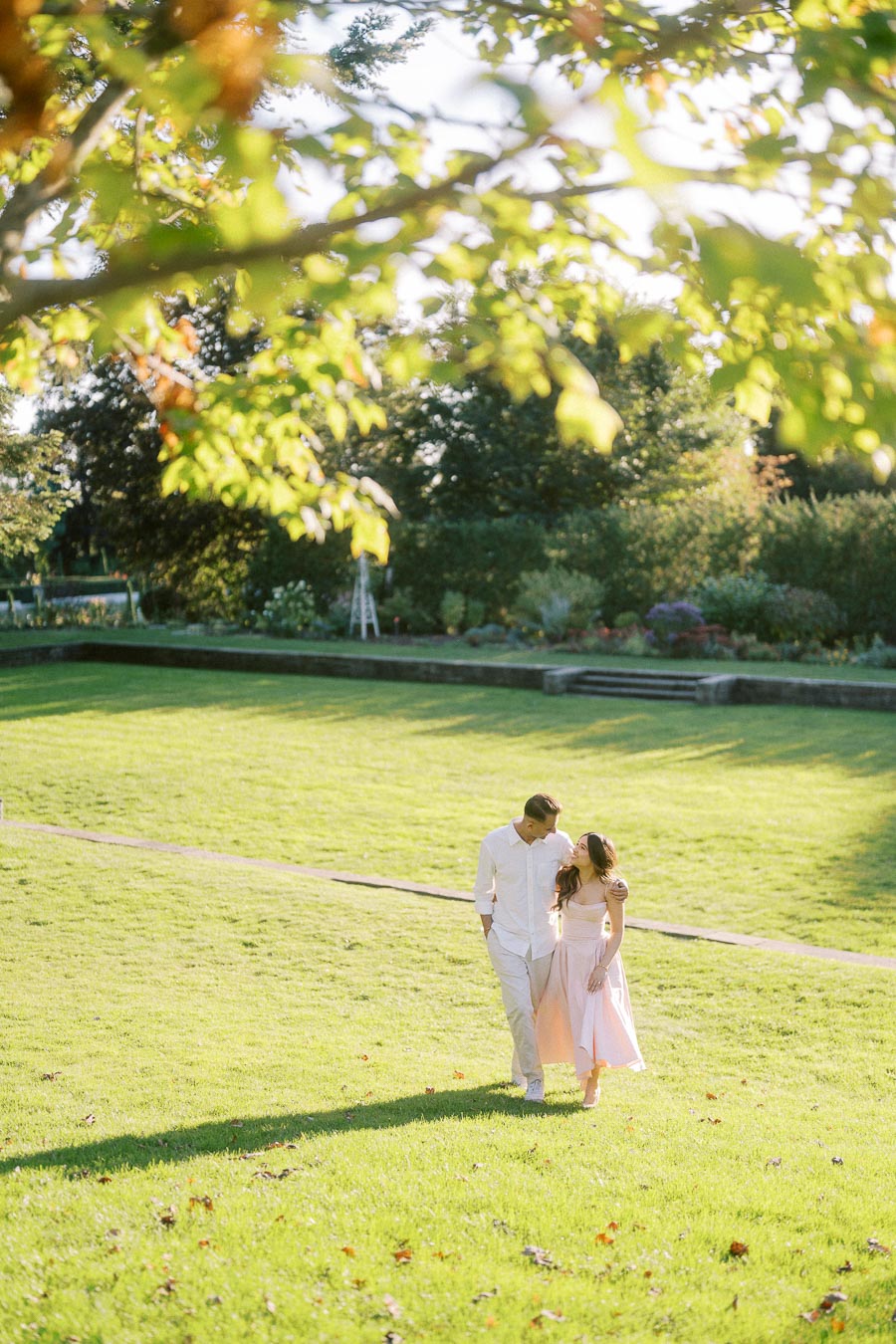 A couple in light-colored clothing walking through a sunny, green park with trees and a garden in the background.