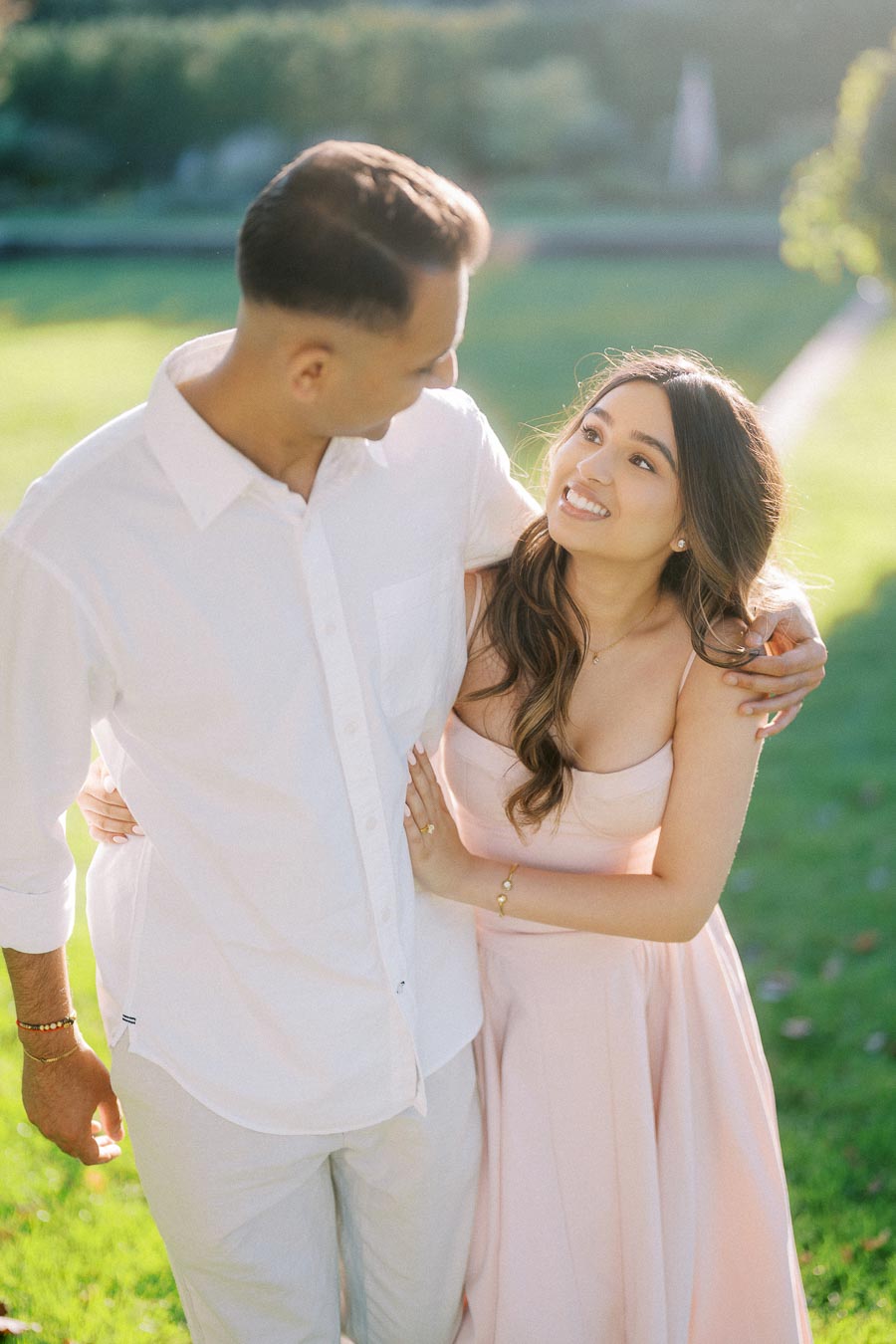 A couple in a sunlit garden, smiling and embracing while wearing elegant attire. The woman in a pink dress looks up at the man in a white shirt, enjoying a romantic moment.