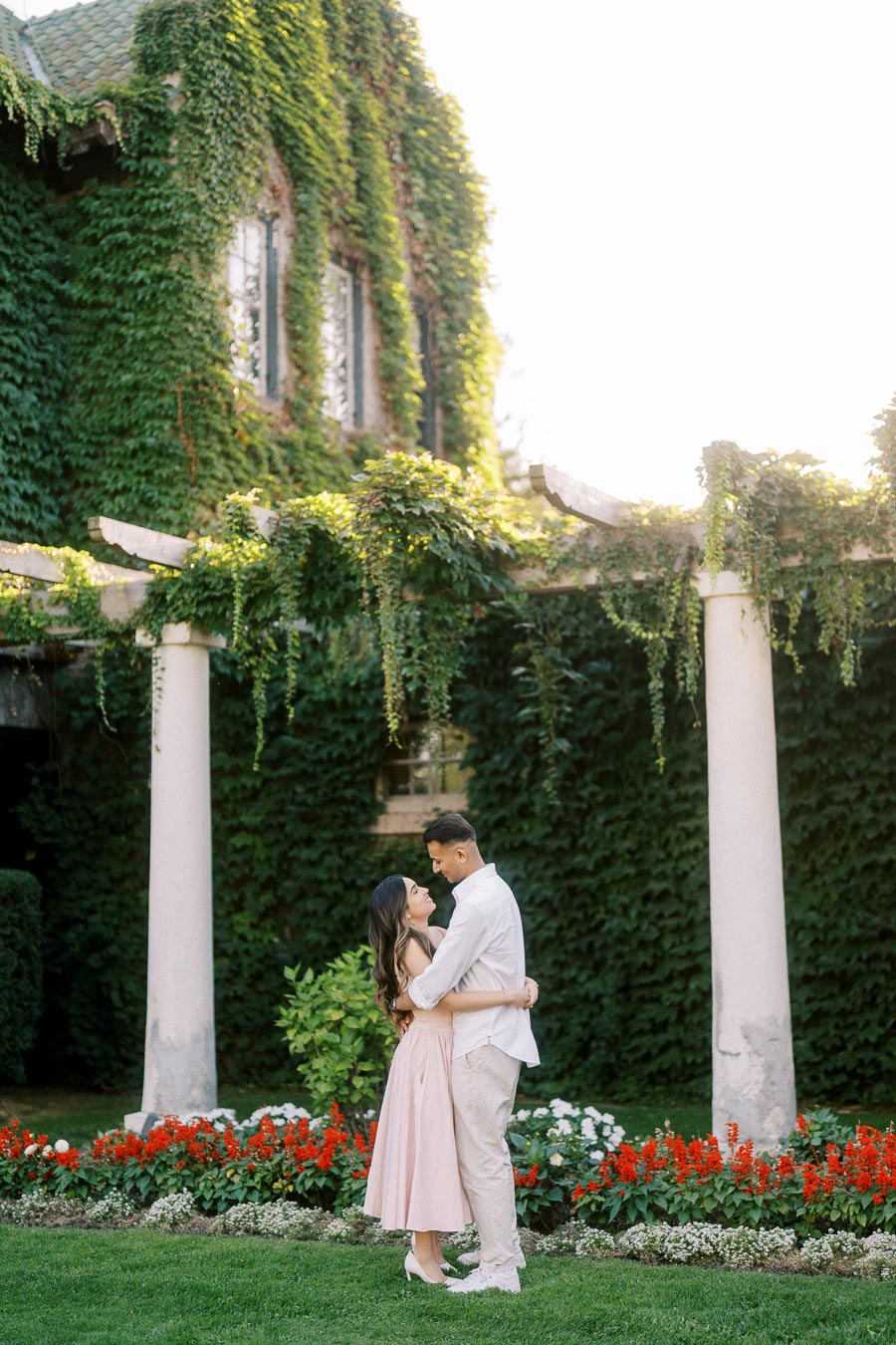 A couple embraces in a lush garden with a vine-covered building backdrop, surrounded by vibrant red and white flowers and green ivy.