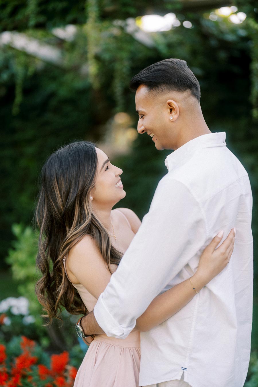 A couple smiling and embracing each other in a garden setting with lush greenery and vibrant flowers in the background.