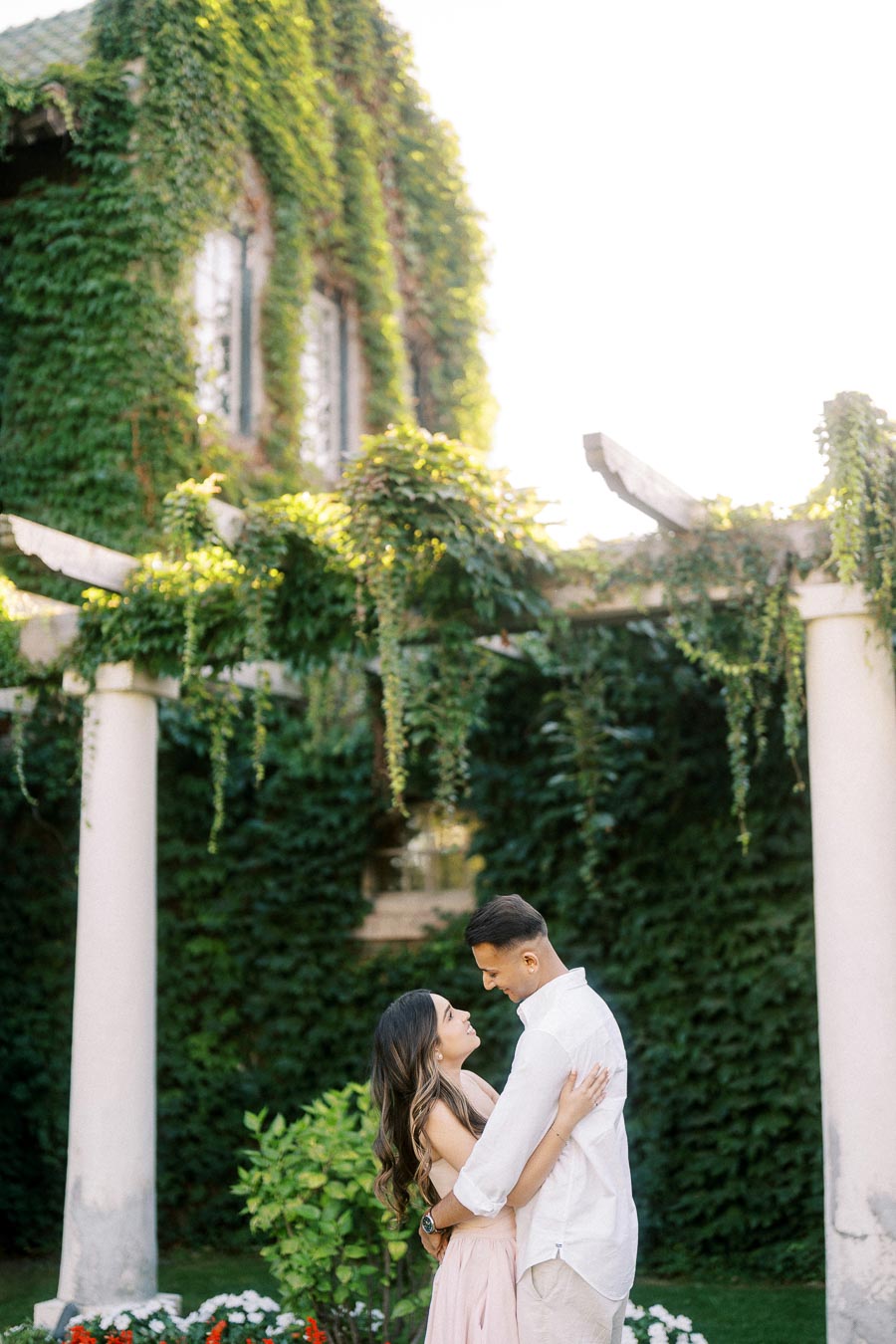 A couple embracing in a lush garden, with a vine-covered building and pergola in the background, conveying romance and serenity.