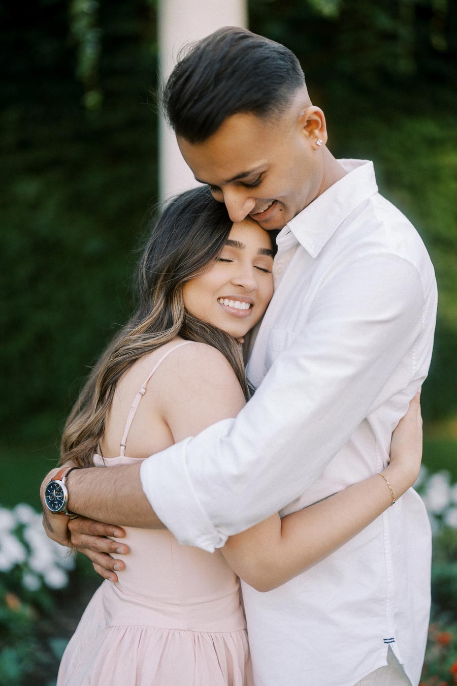 A couple embracing lovingly in a garden setting, with the woman smiling and eyes closed, both wearing light-colored clothing.