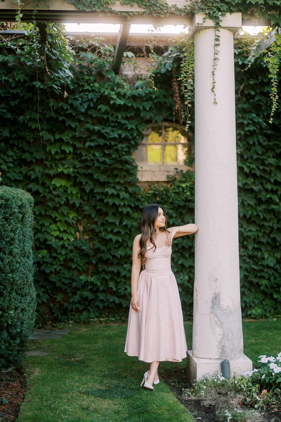 A woman in a pink dress posing against a white column in a garden with lush green ivy on the walls and vibrant plants surrounding the area.