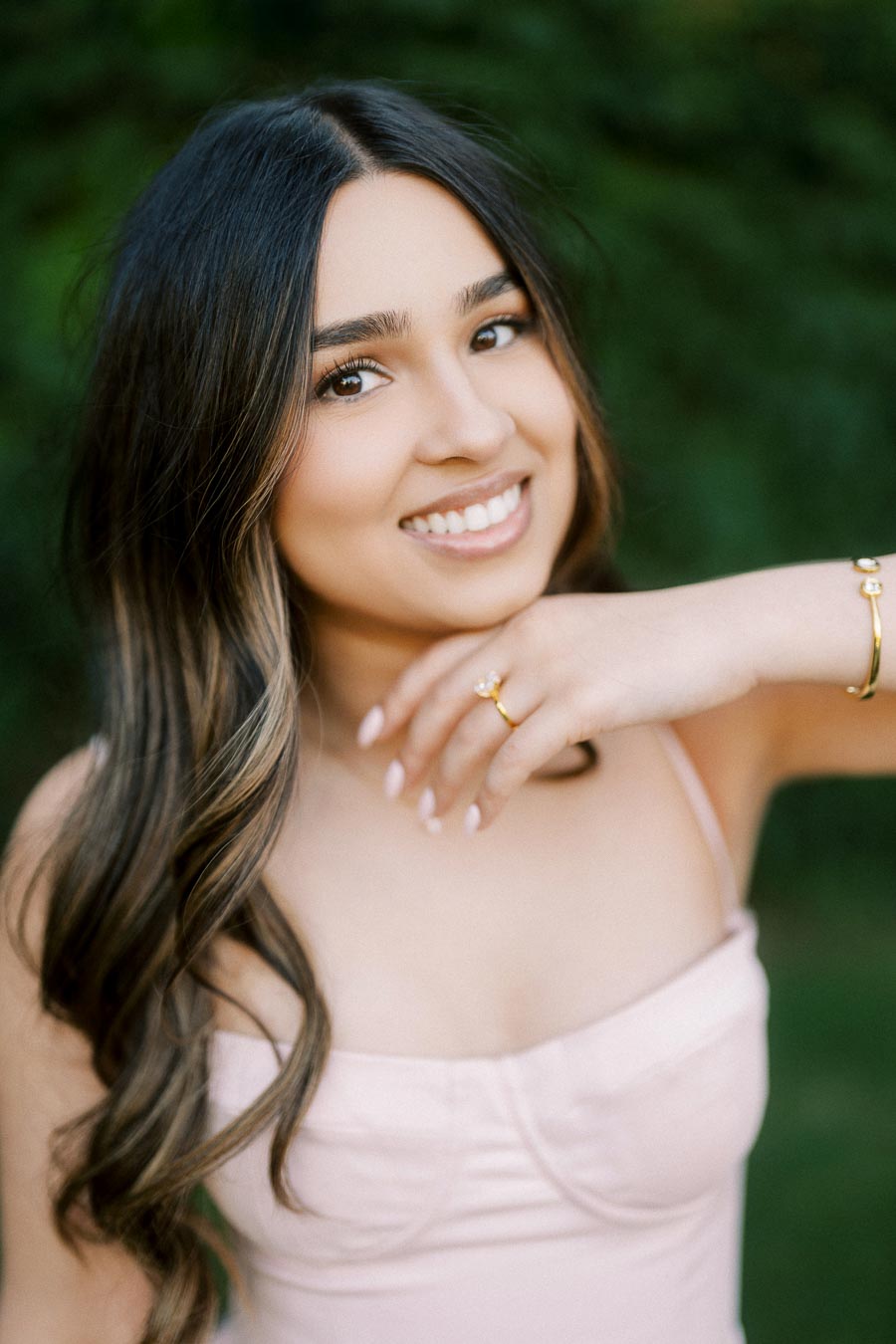 Smiling woman with long, wavy hair wearing a light pink top and gold jewelry, posing outdoors with a green blurred background.