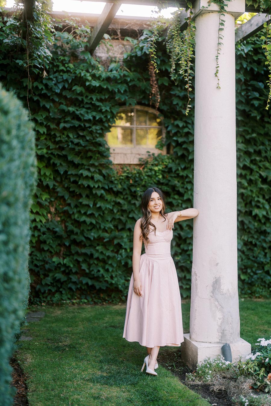 Young woman in a pink dress leaning against a column in a lush garden setting with ivy-covered walls and blooming flowers.