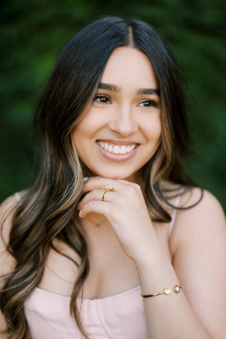 Smiling woman with long dark hair wearing a light pink dress, gold bracelet, and ring, posing outdoors with a blurred green background.