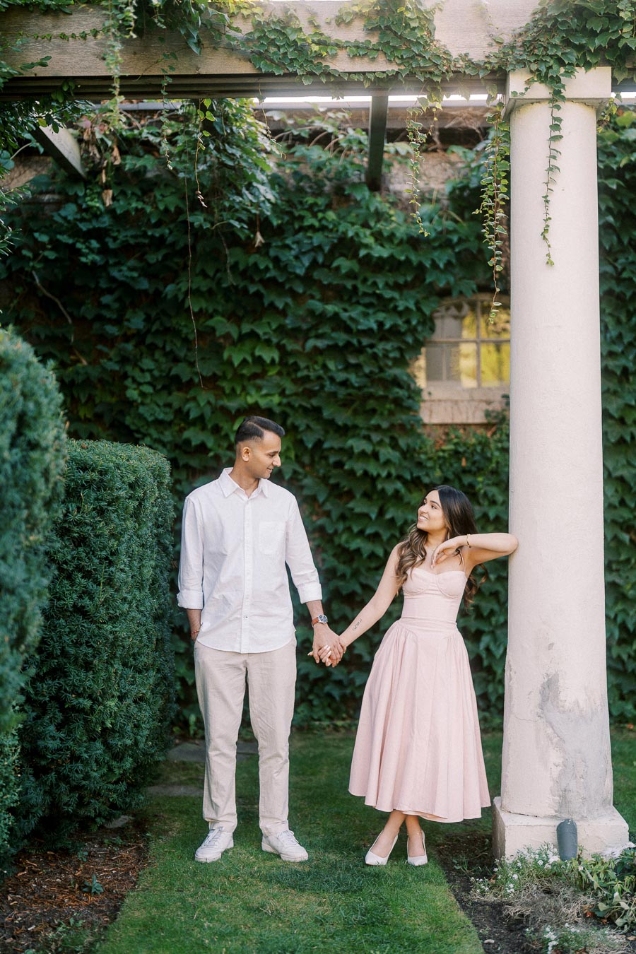 A couple holding hands in front of a lush green ivy-covered wall and columns, with the woman in a pink dress and the man in a white shirt and beige pants, captured in a romantic garden setting.