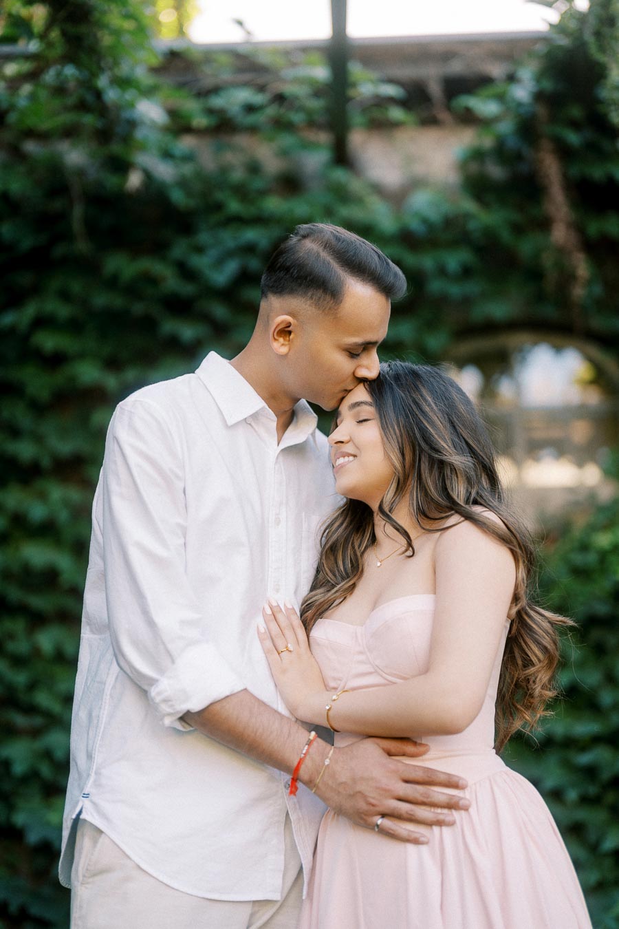 Romantic couple embracing in lush garden setting, with man in white shirt gently kissing woman's forehead, woman wearing a pink dress and smiling serenely.