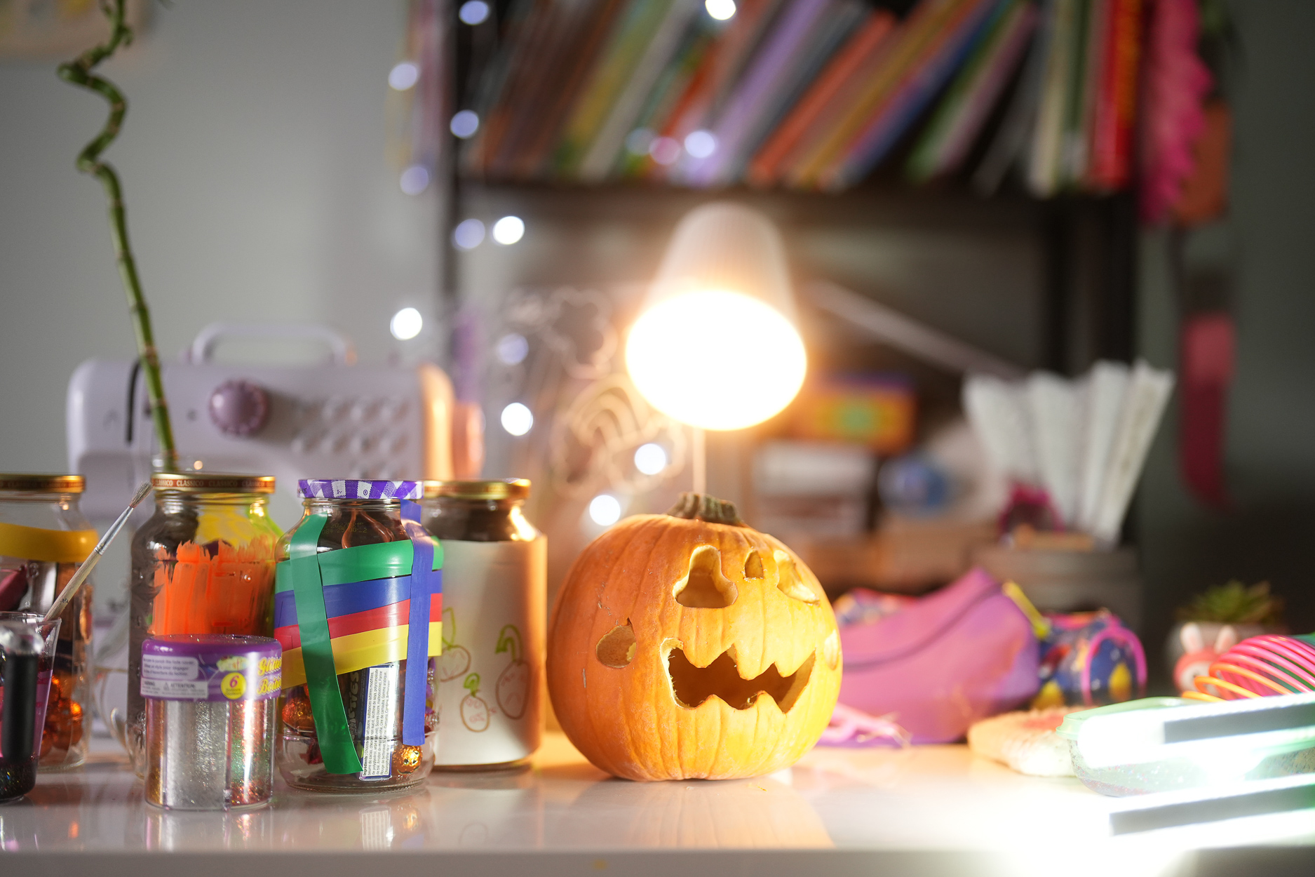 A brightly lit Halloween-themed desk featuring a carved jack-o'-lantern surrounded by colorful craft supplies and jars filled with art materials, with a glowing lamp in the background.