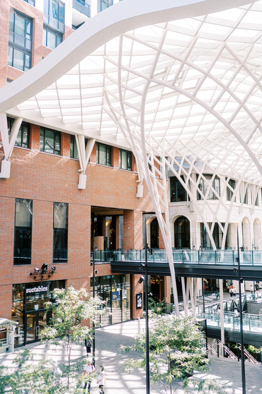 Modern urban shopping area with a distinctive geometric roof, featuring a blend of brick and glass architecture, green trees, and a prominent store entrance.