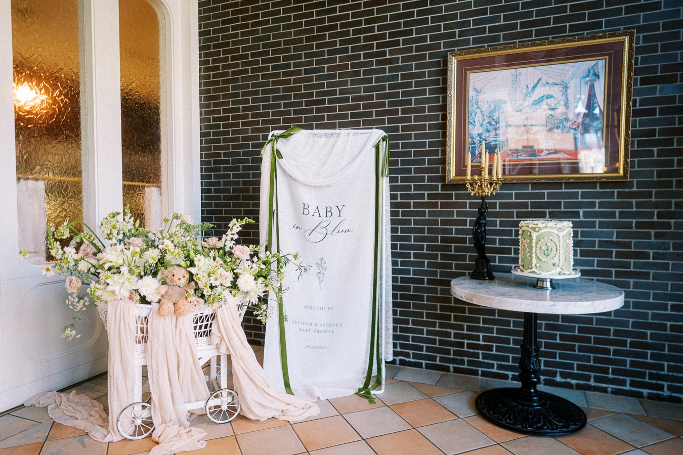 Elegant baby shower setup with floral decorations and a cake. White and green theme with a banner reading Baby in Bloom against a dark brick wall and framed artwork.