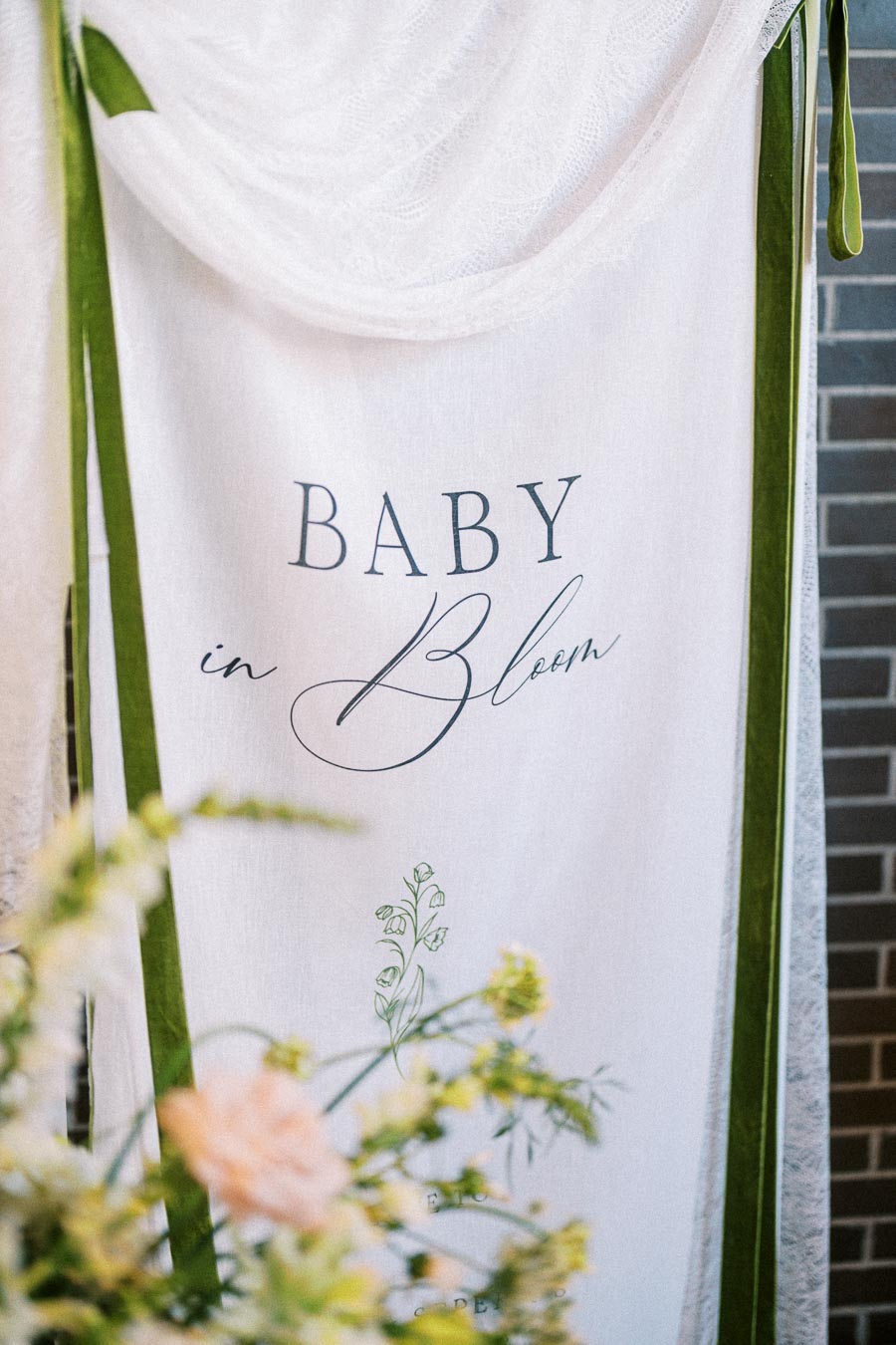Elegant Baby in Bloom banner with floral design, featuring delicate flowers in the foreground and a draped white fabric with green ribbons in a modern setting. Ideal for garden-themed baby showers or botanical-inspired events.