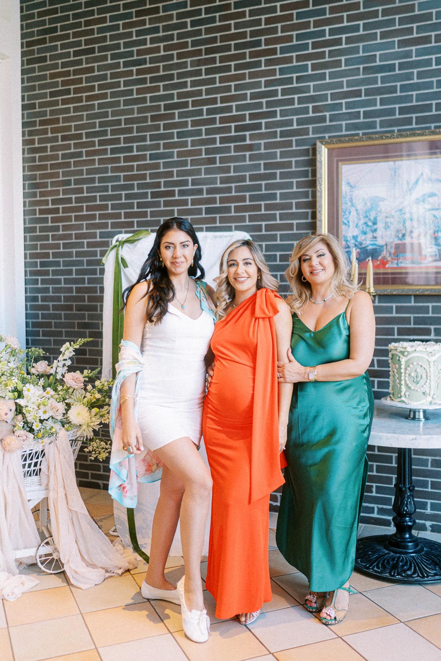 Three women in stylish dresses—one in white, one in orange, and one in green—pose together, standing on tiled flooring in front of a decorative setup with flowers and a cake, against a brick wall background.