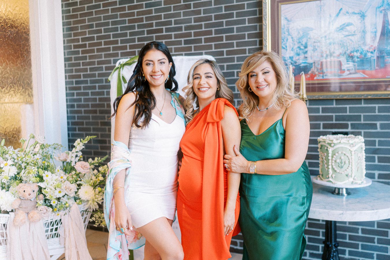 Three women dressed elegantly in vibrant dresses pose together at a celebration, with floral arrangements and a decorative cake in the background.