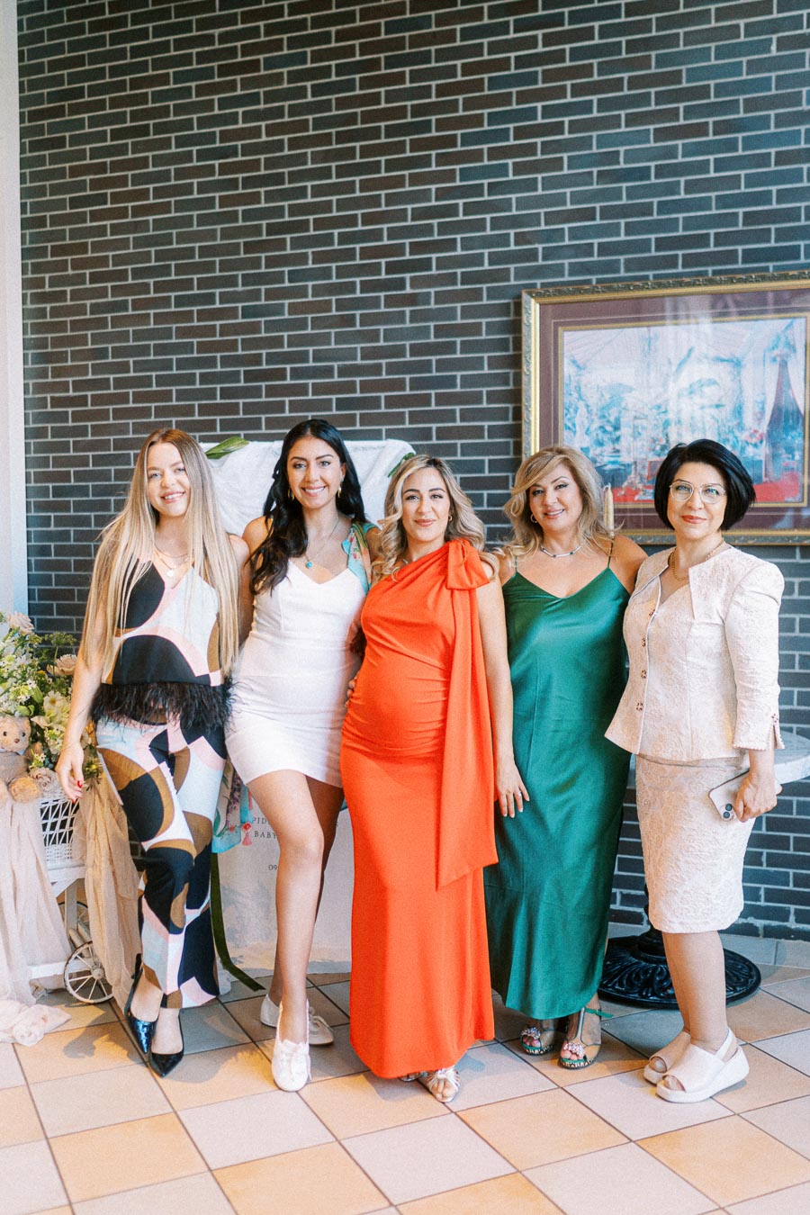 A group of five women smiling and posing together at a social event, each wearing elegant dresses of various styles and colors, standing in front of a brick wall.