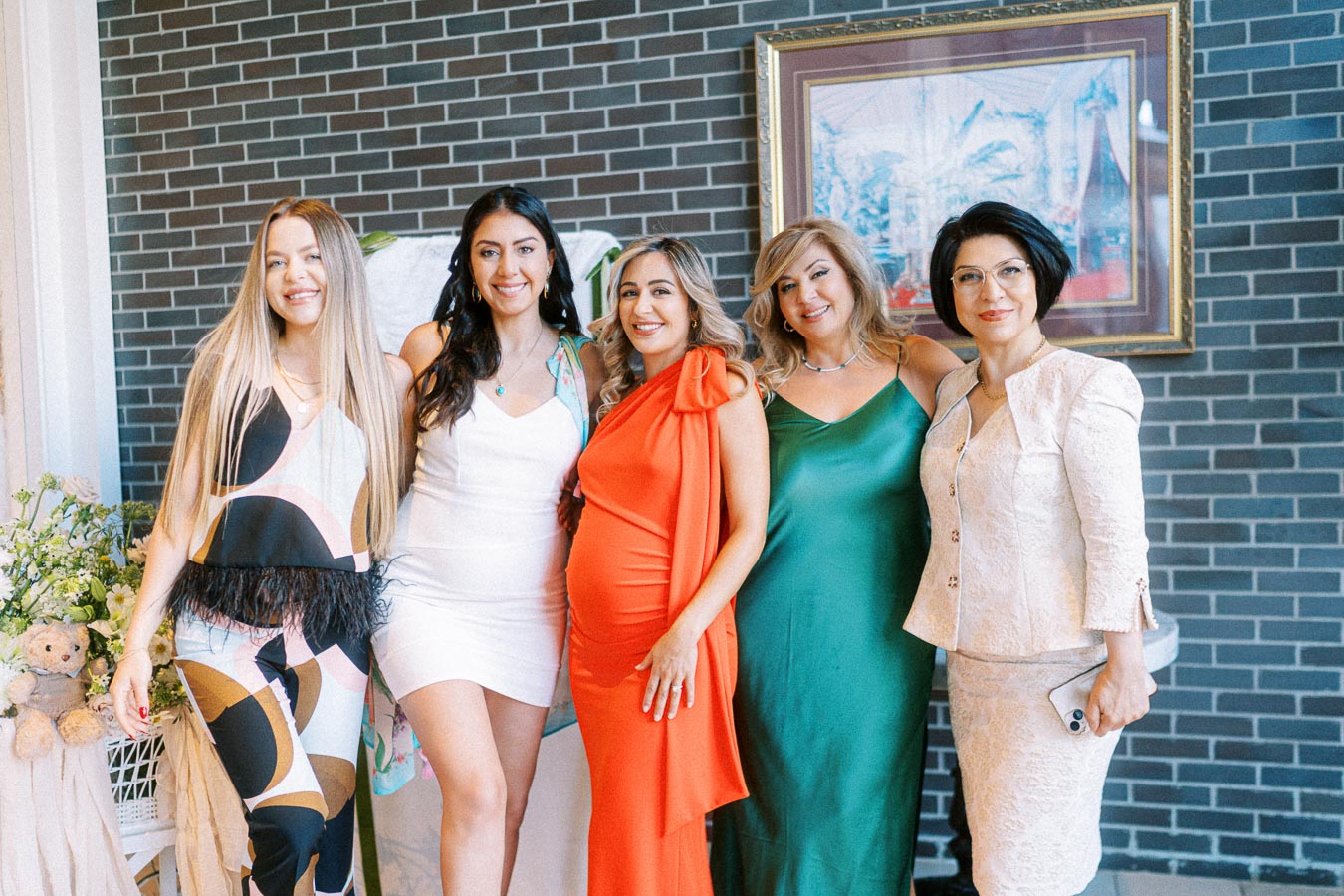A group of five women in elegant dresses posing together at an indoor event, featuring a brick wall and framed artwork in the background.
