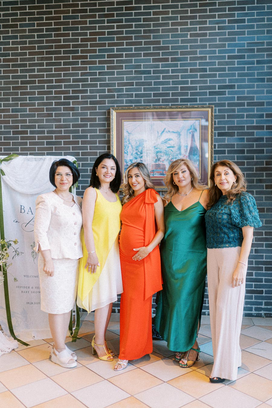 Group of women smiling at a baby shower, standing in front of a brick wall with elegant dresses, including a pregnant woman in a red outfit, celebrating with friends for a special occasion.