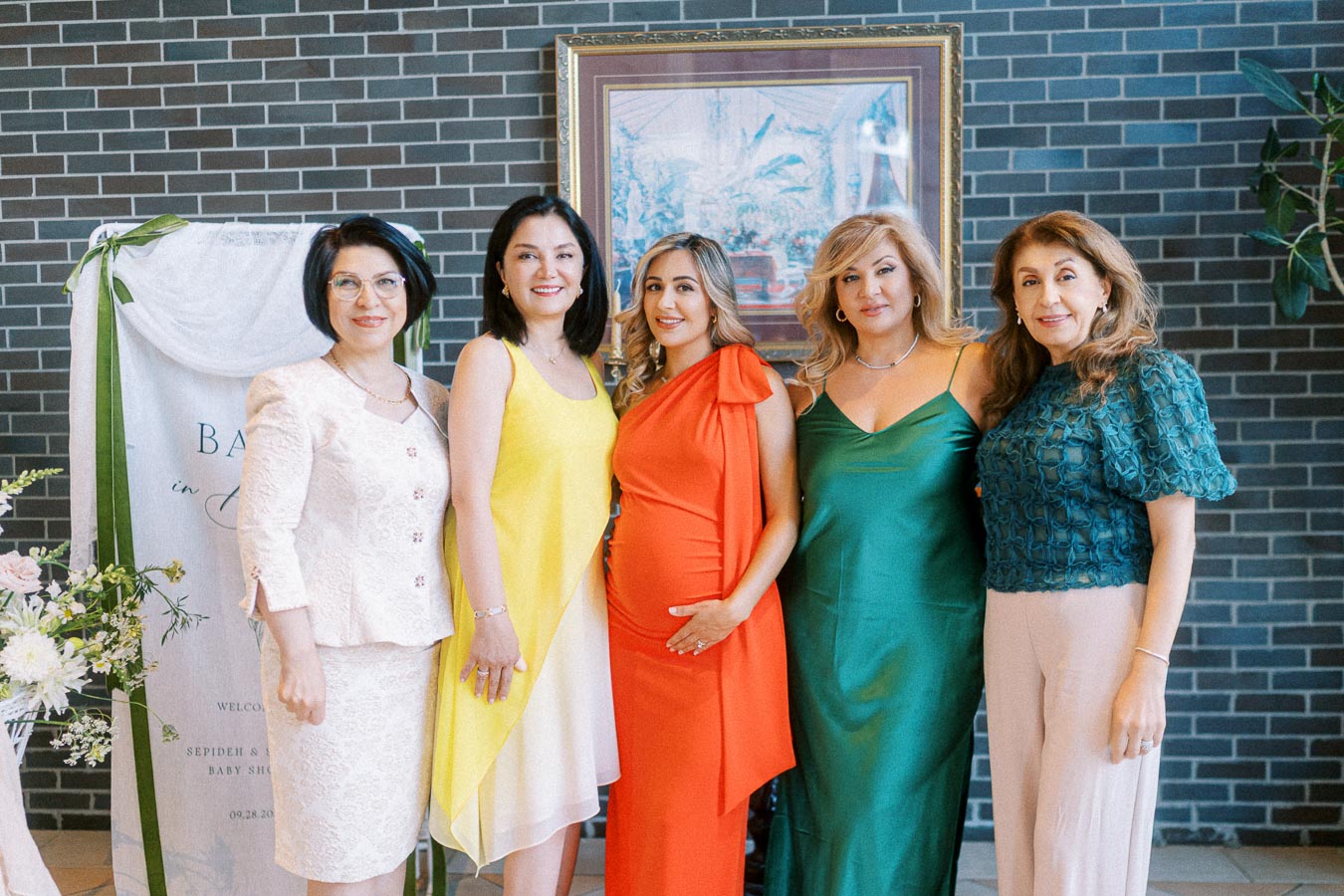 Five women posing together at a baby shower, wearing colorful dresses. A decorative sign and floral arrangements are visible in the background, creating a festive atmosphere.