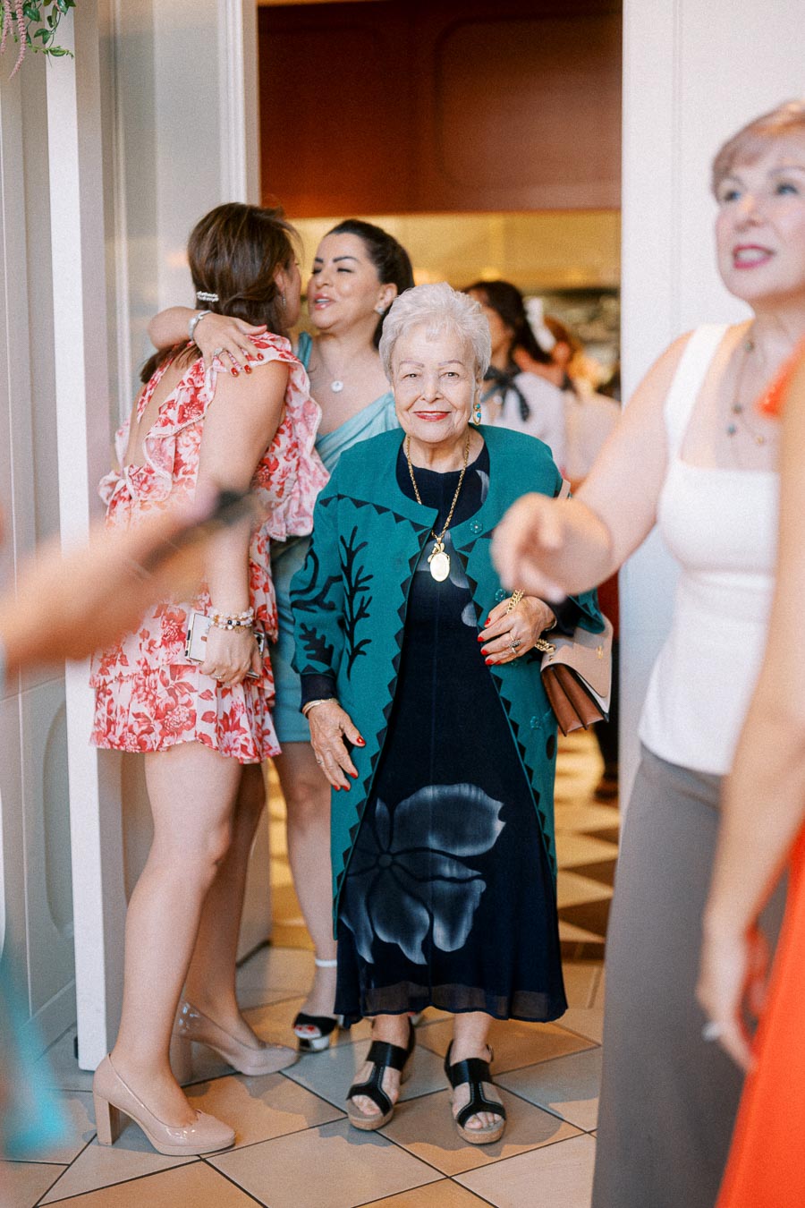 A joyful gathering at a social event, featuring a smiling elderly woman in a stylish blue-green outfit with a floral pattern, surrounded by people greeting each other warmly. The atmosphere is vibrant, with guests dressed in colorful attire, highlighting the convivial and celebratory mood of the occasion.