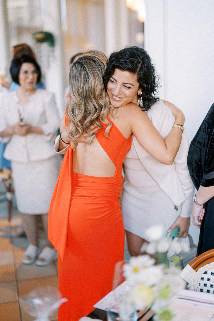 Two women warmly embracing at a social gathering, one wearing a vibrant red dress, the other in a chic white outfit, surrounded by other attendees.