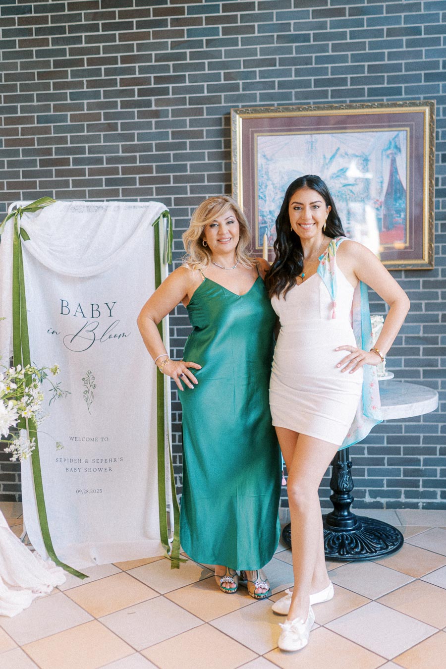 Two women posing at a Baby in Bloom themed baby shower, standing next to a decorative sign with brick wall background.