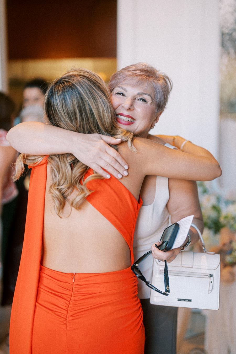 Two women sharing a warm embrace at a gathering, one wearing a vibrant orange dress and the other holding a white handbag and glasses, exuding joy and connection.