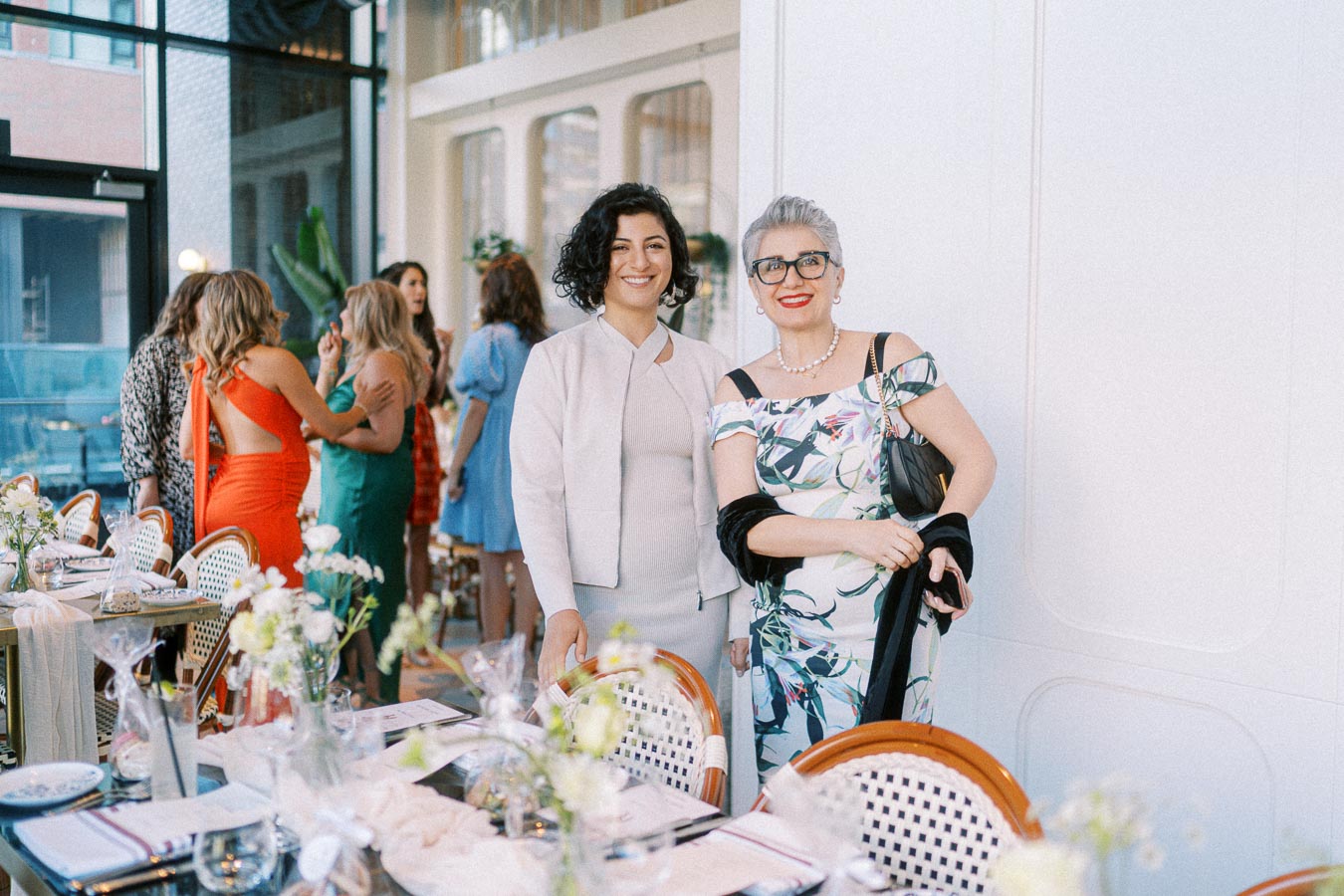 Two women smiling at a stylish indoor event, surrounded by elegantly set tables and other guests in colorful attire, creating a lively and sophisticated atmosphere.