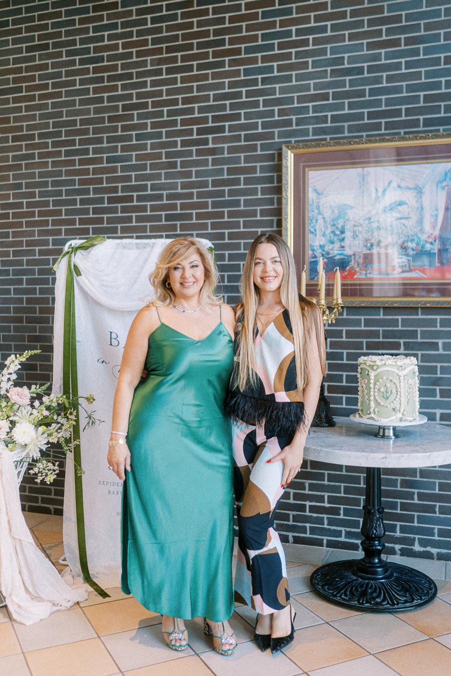 Two women elegantly dressed, smiling at a stylish event with a decorative cake on a marble table and floral arrangements, set against a brick wall backdrop.
