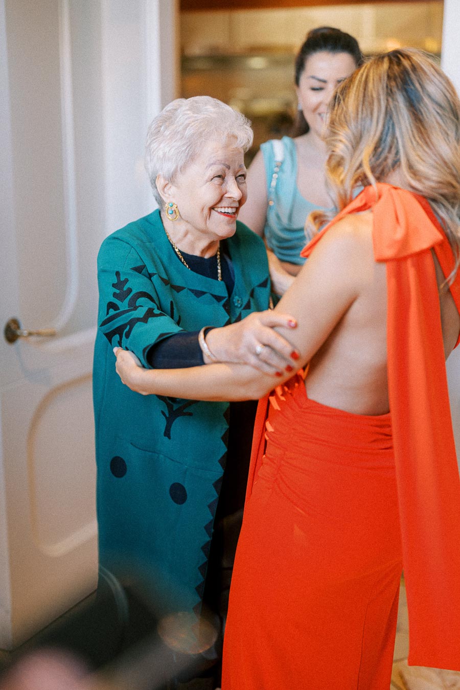 An elderly woman in a teal dress warmly embraces a younger woman in a vibrant red gown, both smiling, with a blurred background suggesting a festive or family gathering.