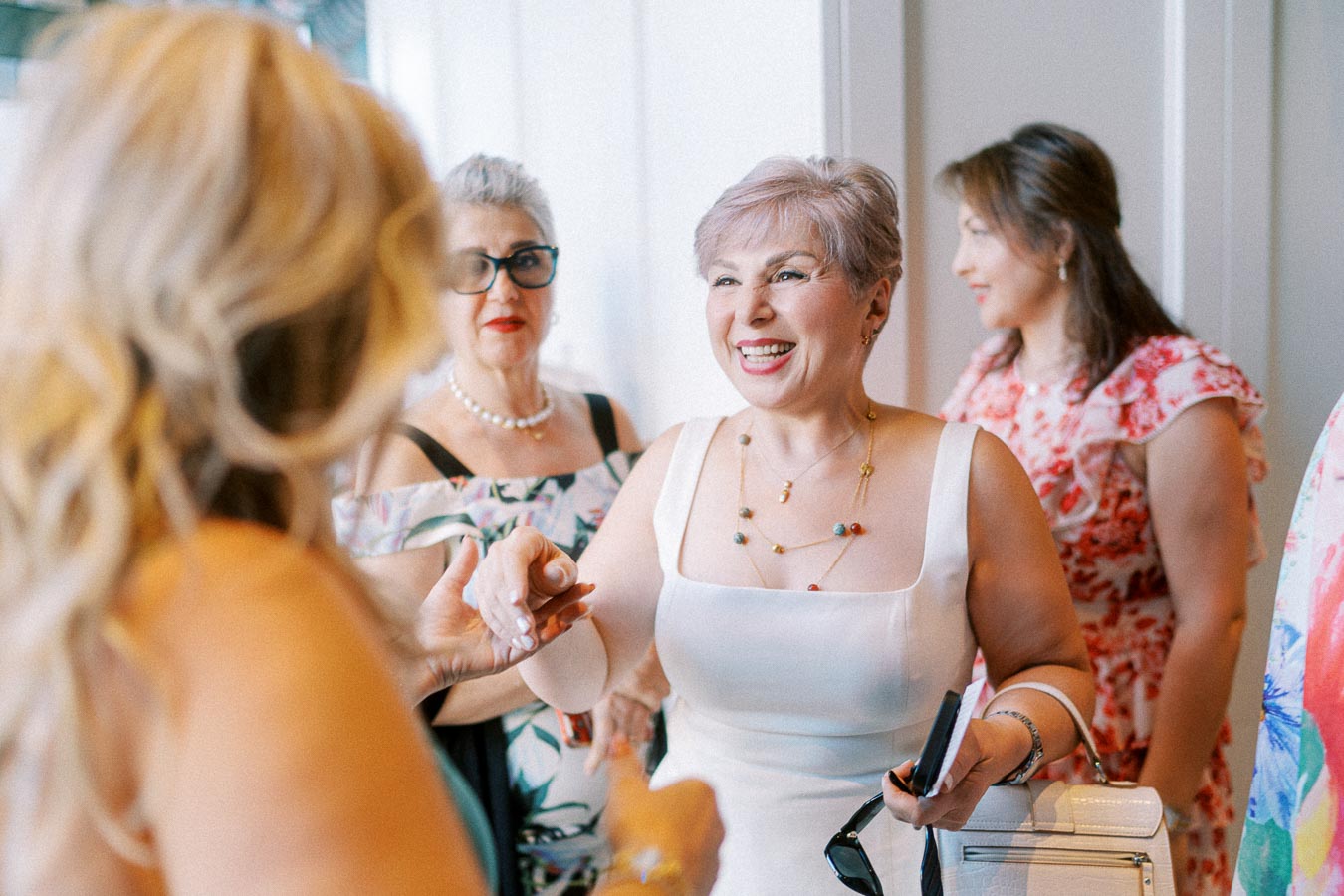 Chatting and smiling women at a social event in a brightly lit room, wearing colorful dresses and accessories.