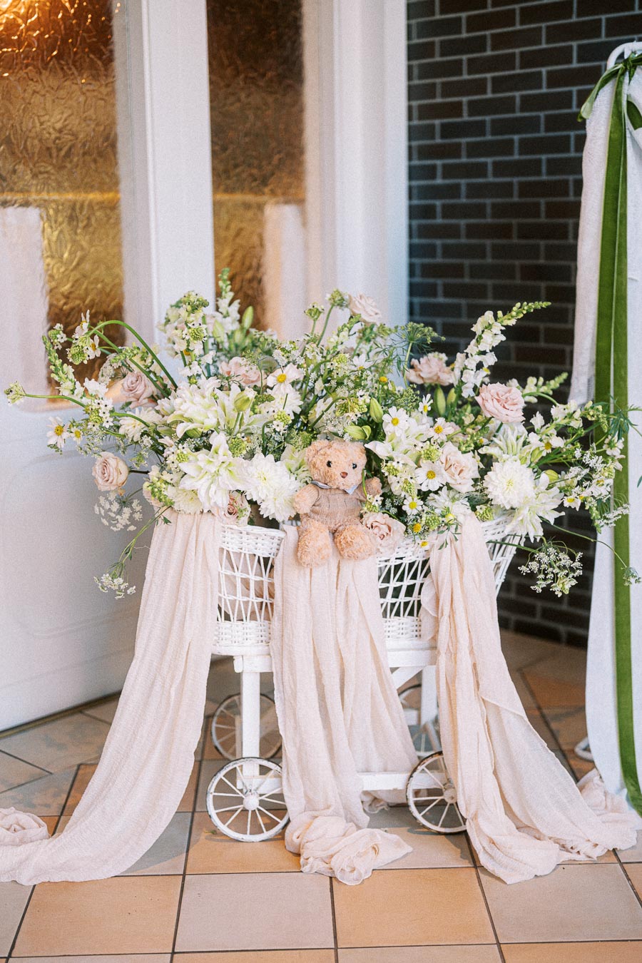 A vintage white wicker pram filled with a vibrant arrangement of white and pink flowers, featuring daisies and roses, draped with soft beige fabric, and a plush teddy bear in the center, creating an elegant and charming decorative display.