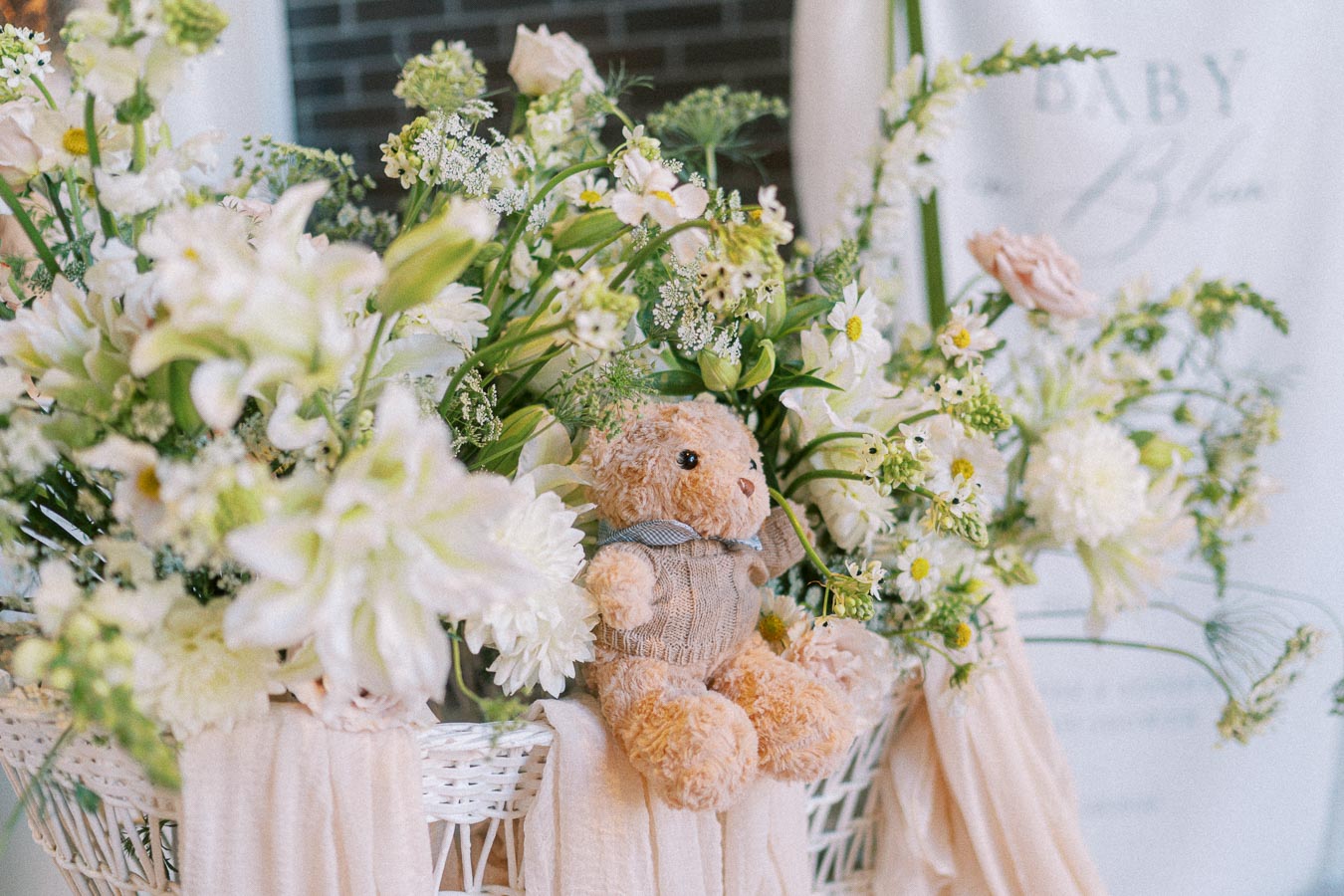 A soft, brown teddy bear in a knitted sweater sits amidst a lush bouquet of white flowers and greenery in a wicker basket, creating a charming and cozy floral arrangement.